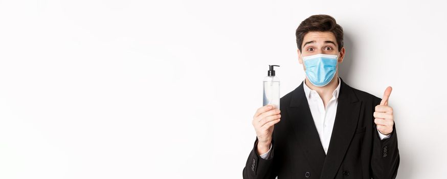 Concept of covid-19, business and social distancing. Close-up of satisfied handsome man in suit and medical mask, showing thumb-up and hand sanitizer, standing against white background.