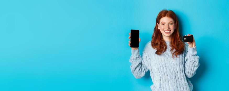 Excited redhead girl showing mobile phone screen and credit card, demonstrating online store or application, standing over blue background.