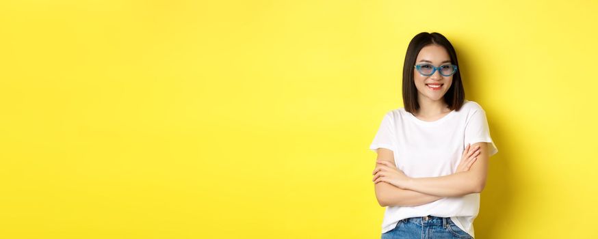 Fashion and lifestyle concept. Sassy and confident asian woman in trendy sunglasses looking self-assured at camera, standing over yellow background.