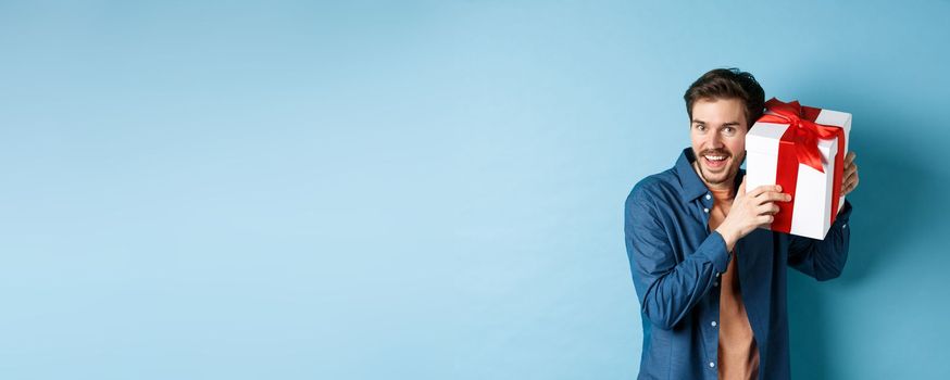 Valentines day. Happy young man got present on special holiday, trying guess what inside gift box and smiling, standing over blue background.