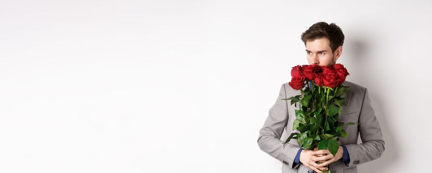 Romantic man in suit smell bouquet of roses and looking pensive, standing over white background. Concept of valentines day.