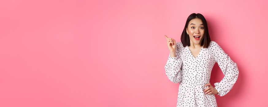 Cute asian woman in dress pointing upper left corner copy space. Korean model showing advertisement, standing over pink background.