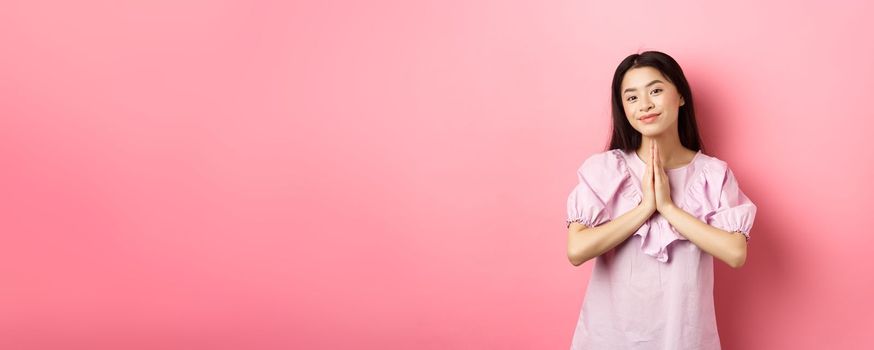 Cute asian girl say thank you, smiling and looking happy, showing namaste gesture in gratitude, standing in dress against pink background.