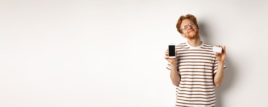 Shopping and finance concept. Pleased young man with red hair smiling from satisfaction, showing smartphone blank screen and credit card, white background.