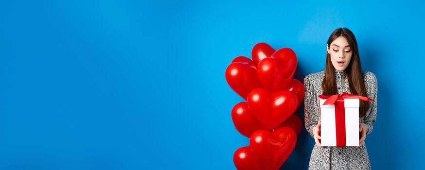 Valentines day. Image of beautiful girl looking surprised at gift box, receive present from lover, standing in dress on blue background.