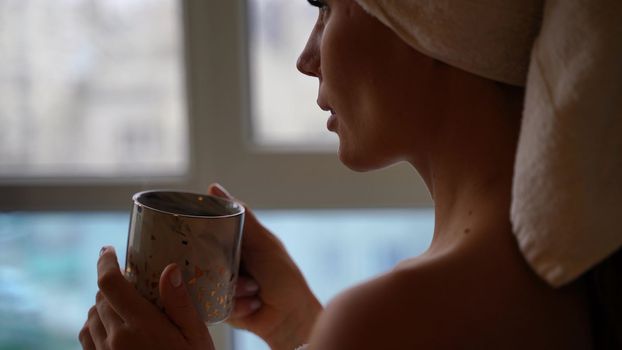 Middle aged woman looks good with bare shoulders in a white towel on her head holds a cup and drinks coffee or tea against the window.