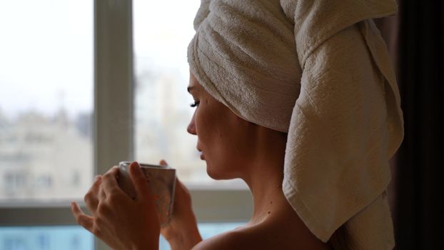 Middle aged woman looks good with bare shoulders in a white towel on her head holds a cup and drinks coffee or tea against the window.
