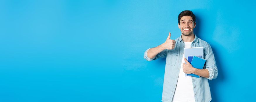 Happy young male model holding notebooks and showing thumb-up pleased, smiling and recommending courses, standing over blue background.