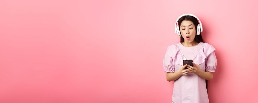 Excited asian girl looking at smartphone screen amused, say wow, listening music in wireless headphones, standing against pink background.