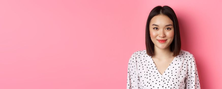 Close-up of cute asian female model looking at camera, blushing and smiling kawaii, standing over pink background.