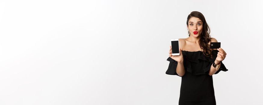 Fashion and online shopping concept. Happy young woman in black dress, showing credit card and mobile screen, standing over white background.