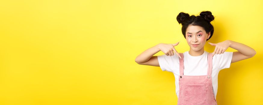 Stylish sassy girl smiling pleased, hinting on good deal, pointing fingers down at promo offer, standing in summer clothes against yellow background.
