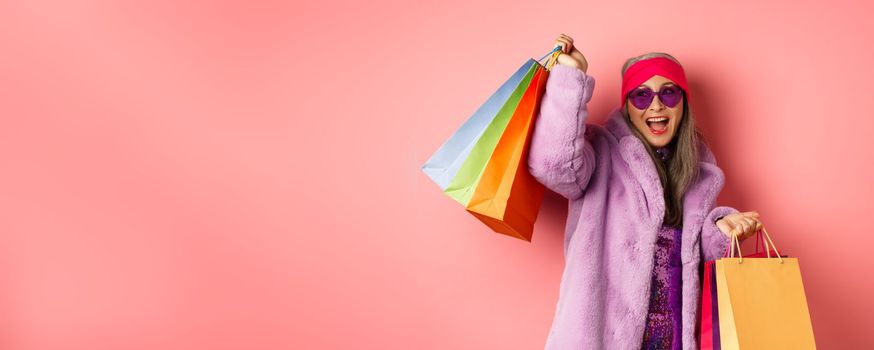 Stylish asian senior woman going shopping, wearing trendy clothes and sunglasses, holding store bags and dancing happy, pink background.