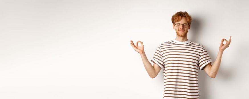 Calm and relaxed young man with red messy hair, spread hands sideways in mudra gesture and smiling, practice yoga or meditating, white background.