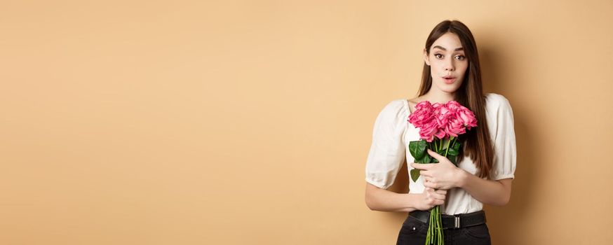Valentines day. Image of surprised girlfriend thanking for flowers, receive pink roses from lover and looking grateful at lover, standing on beige background.
