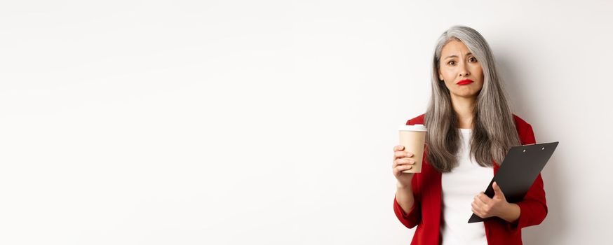 Tired and disappointed asian businesswoman with grey hair, drinking coffee in paper cup and looking gloomy at camera, standing over white background.