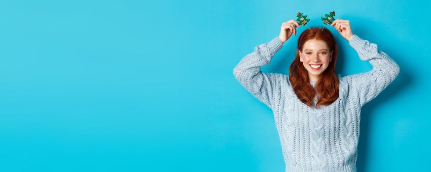 Winter holidays and Christmas sales concept. Beautiful redhead female model celebrating New Year, wearing funny party headband and sweater, smiling at camera.