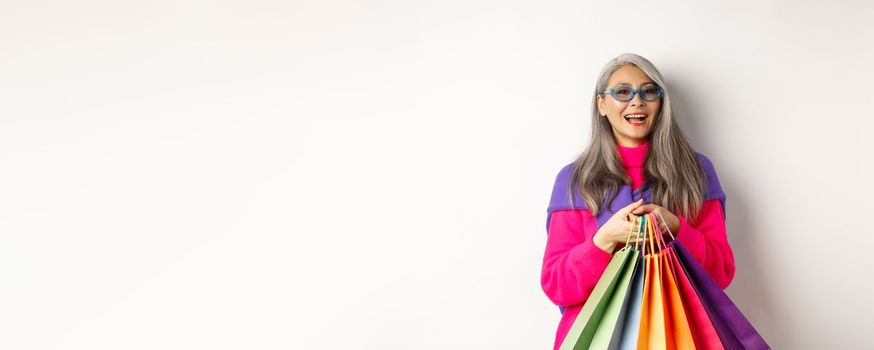 Stylish senior asian woman in sunglasses going shopping on holiday sale, holding paper bags and smiling, standing over white background.