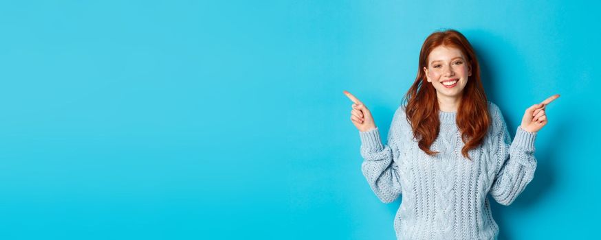 Winter holidays and people concept. Cute teen girl with red hair, smiling and pointing fingers sideways, showing advertisements, standing over blue background.