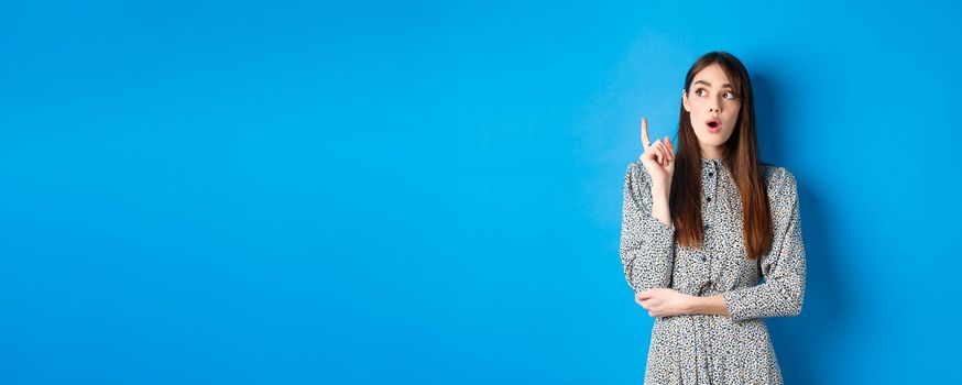 Thoughtful attractive girl in dress pitching an idea, raising finger and looking up, have a plan, standing on blue background.