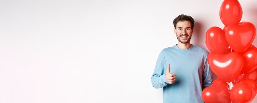 Valentines day and lovers concept. Happy young man showing thumbs up as standing with red hearts balloon, bring romantic gift on date, standing over white background.