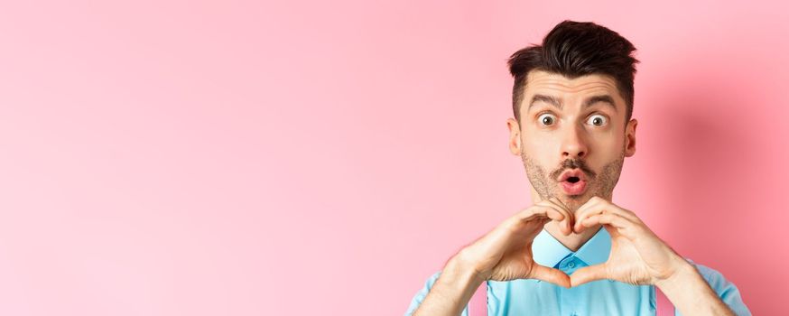 Valentines day concept. Close up of handsome caucasian man falling in love, looking amazed and showing heart gesture, pink background.