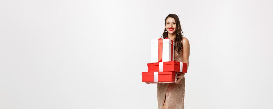 Christmas party and celebration concept. Full-length of stylish woman with red lips, glamour dress, holding gifts and smiling happy, standing over white background.