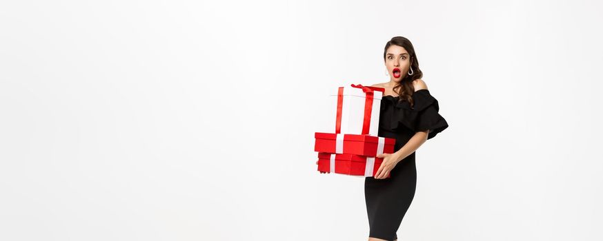 Full length of attractive female model in glamour dress, red lips, holding christmas gifts and looking amazed, receive presents, white background.