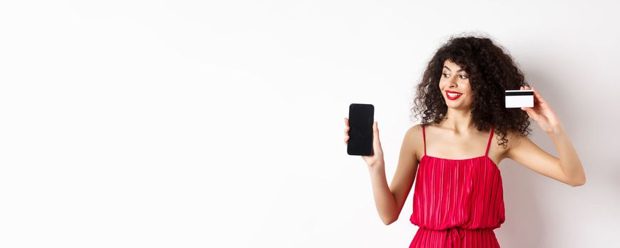 Online shopping concept. Elegant curly-haired woman in red dress showing plastic credit card and empty mobile phone screen, standing over white background.