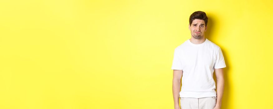 Disappointed guy looking upset, sulking and frowning, standing displeased against yellow background.