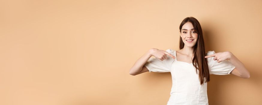 Smiling woman pointing at center, showing logo with happy face, standing in white dress on beige background.