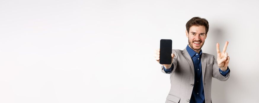 E-commerce and online shopping concept. Cheerful man in business suit showing v-sign and empty mobile phone screen, demonstrate smartphone app, white background.