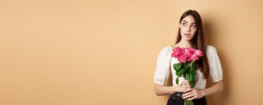 Romantic Valentines day concept. Dreamy woman with bouquet of pink rose, looking aside at logo, holding flowers on beige background.