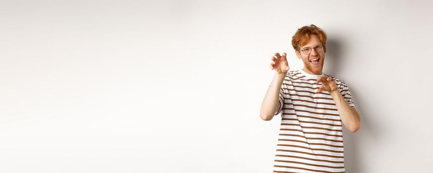 Funny young man with red hair and beard showing claws gesture and roaring, having fun, standing happy over white background.