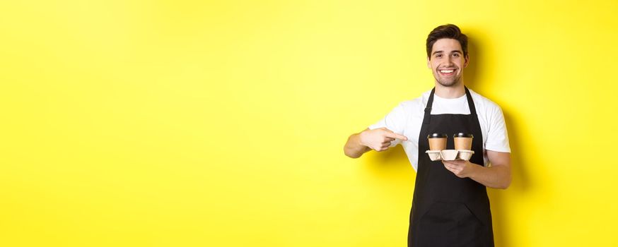 Handsome barista holding two cups of takeaway coffee, pointing finger at drinks and smiling, standing in black apron against yellow background.