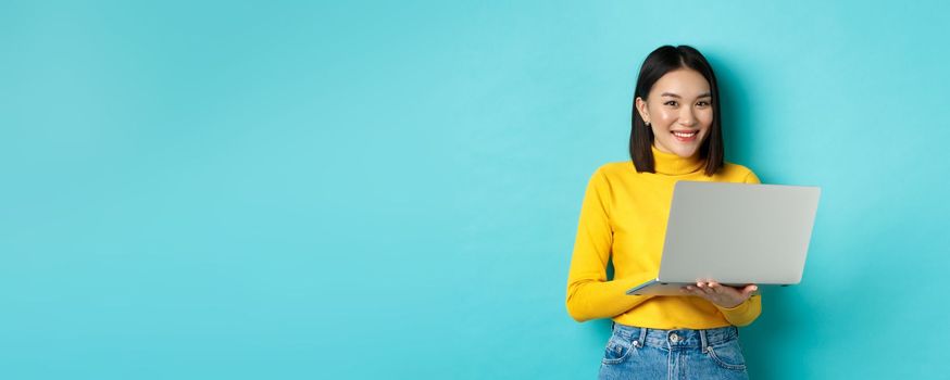 Happy asian woman in yellow pullover using laptop, shopping online or working, standing over blue background.
