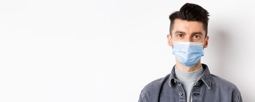 Pandemic lifestyle, healthcare and medicine concept. Close up of candid young man wearing medical mask from covid-19, taking care of health, standing on white background.