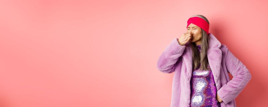 Portrait of stylish old asian woman in fashionable purple coat and dress, shut nose from bad smell, express disgust and aversion as something sink awfully, standing over pink background.