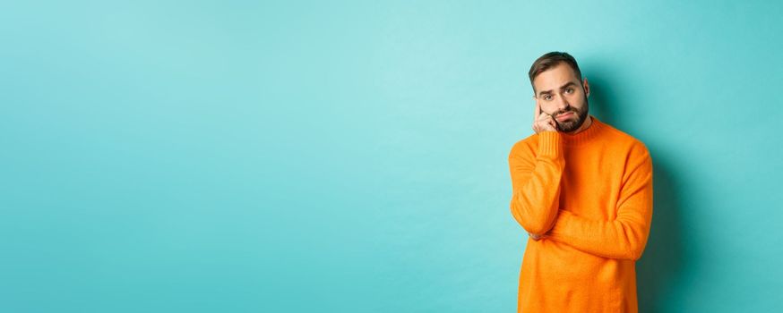 Sad and gloomy man with beard, looking indesicive and sad, thinking about something and sulking, standing over turquoise background.