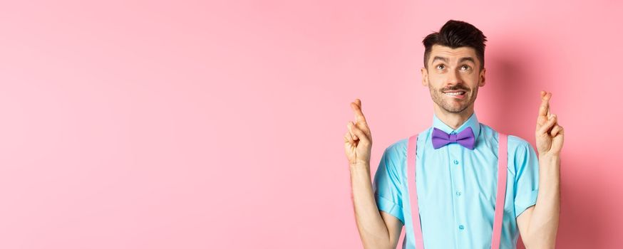 Smiling young man in bow-tie, making wish, praying and looking up, cross fingers for good luck, standing hopeful over pink background. Copy space