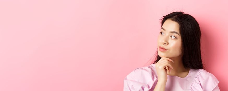 Close-up portrait of romantic asian girl looking aside at logo, thinking or daydreaming, standing against pink background.