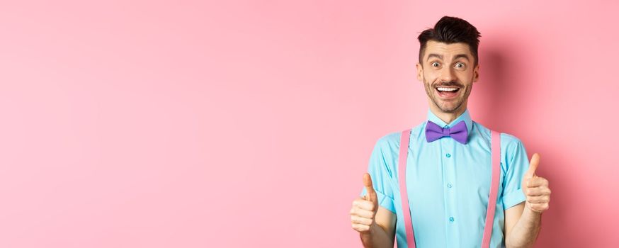 Excited caucasian guy showing thumbs up and praising great work, well done gesture, standing in suspenders and bow-tie on pink background.