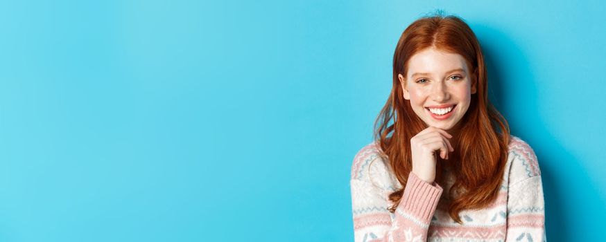 Close-up of beautiful redhead girl smiling, having conversation and staring at camera interested, standing over blue background.