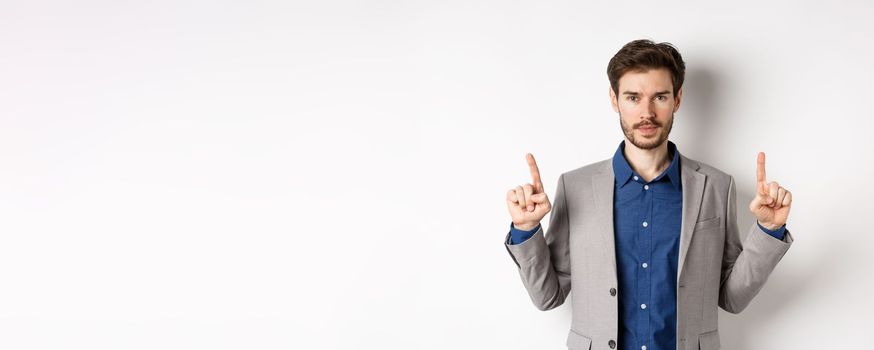 Serious businessman with beard wearing suit, pointing fingers up, look here gesture, advertising on white background.