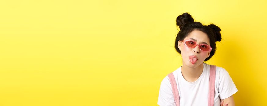 Summer and beauty concept. Childish asian girl in sunglasses showing tongue, being rude, standing on yellow background.