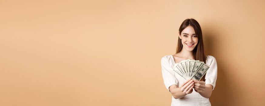 Young woman showing dollar bills and smiling, standing with money on beige background. Concept of loan and insurance.