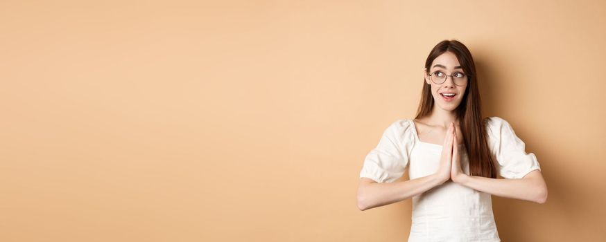 Hopeful girl holding hands in pray, begging for dream come true, looking aside excited, standing on beige background. Copy space