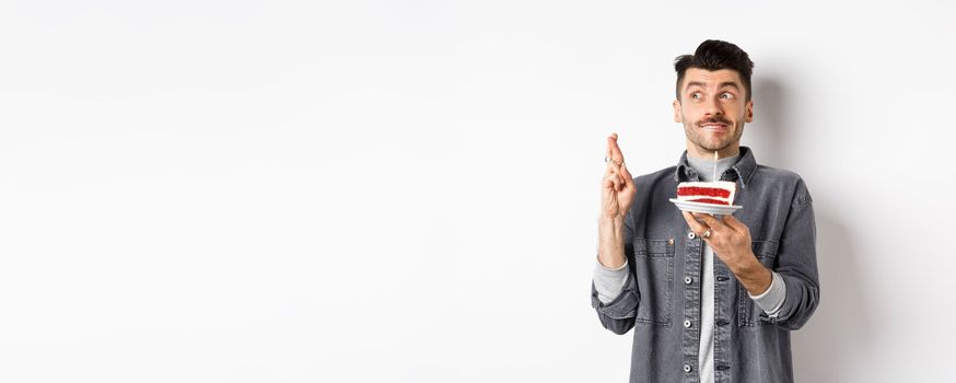 Birthday. Happy young man celebrating, making wish with cake and cross fingers, looking hopeful aside, standing on white background.