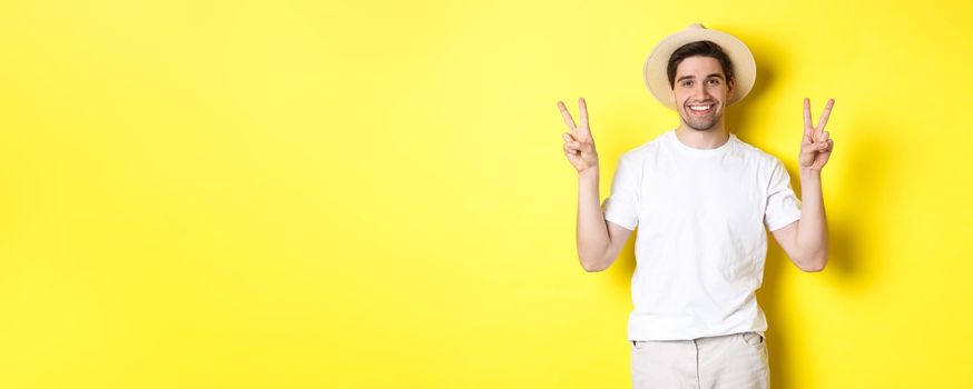 Concept of tourism and vacation. Happy male tourist posing for photo with peace signs, smiling excited, standing against yellow background.