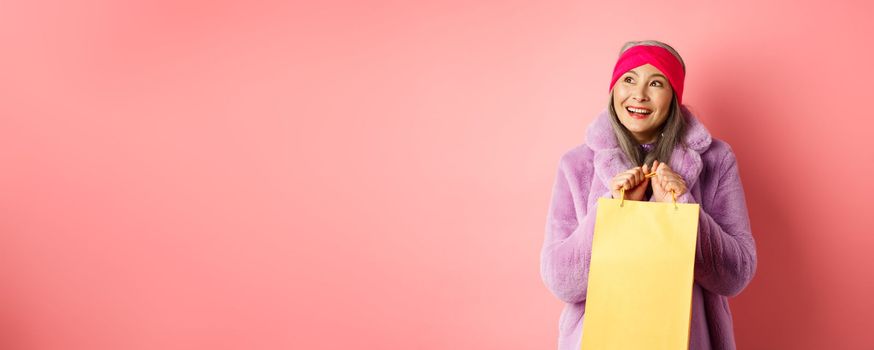 Shopping and fashion concept. Beautiful asian senior woman looking dreamy at upper left corner and imaging something, holding yellow paper bag with gift, pink background.